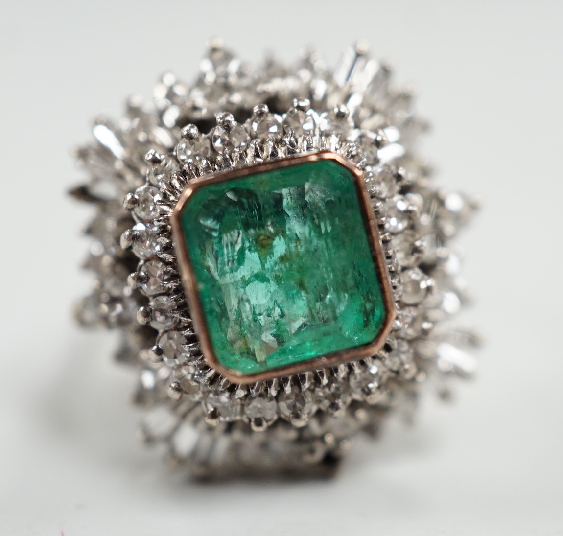 A white metal and single stone emerald set dress ring, bordered with two stepped bands of round and baguette cut diamonds, size K, gross weight 7.6 grams.